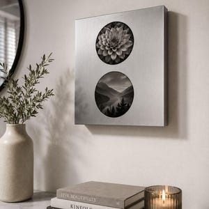 May include: A rectangular brushed silver metal wall art piece featuring two circular cutouts. The top circle displays a black and white dahlia flower, while the bottom circle shows a black and white landscape of mountains, a lake, and pine trees.