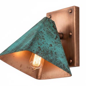 May include: A wall-mounted light fixture featuring a rectangular copper backplate with four rivets and a triangular, pyramid-shaped shade. The exterior of the shade has a weathered, teal-green patina finish, while the interior is smooth copper.
