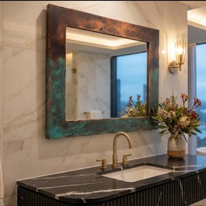 May include: A rectangular wall mirror featuring a wide, textured frame with a weathered copper finish that transitions from dark brown at the top to a vibrant oxidized teal green at the bottom, reflecting a modern bathroom interior with marble walls.