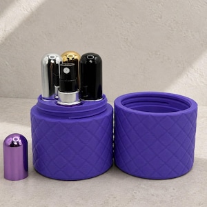 May include: A purple cylindrical container with a quilted texture, featuring a matching lid and a removable insert holding three small spray bottles with silver, gold, and black caps, plus one separate purple cap placed to the left.