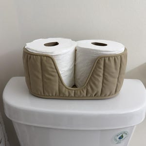 May include: A beige, quilted fabric toilet paper holder designed to hold two standard rolls of white toilet paper. The holder features a U-shaped cutout on the front and a structured, oval-shaped base, providing a soft, padded exterior.