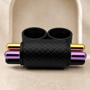 May include: A black cylindrical holder with a diamond-patterned texture, featuring two circular openings at the top. Three horizontal metal rods in gold, purple, and black are inserted through the sides of the central black component.