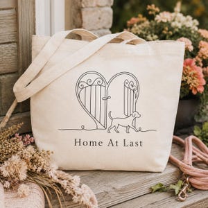 Puede incluir: Un bolso de tela color crema con una ilustración de líneas negras de un perro caminando hacia una puerta abierta en forma de corazón. Debajo del gráfico, el texto Home At Last está impreso en una fuente serif oscura y sencilla. Tiene dos asas.