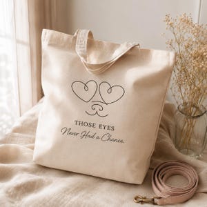 May include: A beige canvas tote bag featuring a black line art graphic of two heart-shaped eyes and a small nose. Below the graphic, the text reads: THOSE EYES Never Had a Chance. The bag has two top handles and is constructed from a light-coloured fabric.