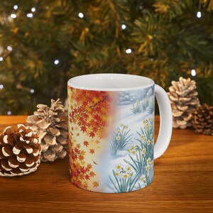 May include: A white ceramic mug featuring a split-design graphic. One side displays vibrant red and orange autumn maple leaves, while the other side depicts white daffodils blooming through a light dusting of winter snow on the ground.