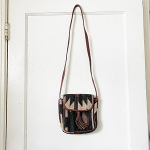 K&ouml;nnte beinhalten: Eine kleine braune Leder-Umh&auml;ngetasche mit einem geometrischen Muster in Schwarz, Wei&szlig; und Braun. Die Tasche hat einen Klappenverschluss und einen Quasten.