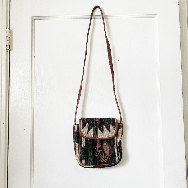 K&ouml;nnte beinhalten: Eine kleine braune Leder-Umh&auml;ngetasche mit einem geometrischen Muster in Schwarz, Wei&szlig; und Braun. Die Tasche hat einen Klappenverschluss und einen Quasten.