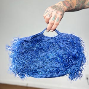 May include: A vibrant blue handbag with a unique, textured design. The bag features a woven base and is covered in looped, fringe-like strands, creating a playful and eye-catching accessory. The bag has two handles.