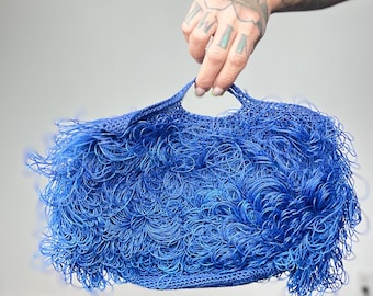 May include: A vibrant blue handbag with a unique, textured design. The bag features a woven base and is covered in looped, fringe-like strands, creating a playful and eye-catching accessory. The bag has two handles.