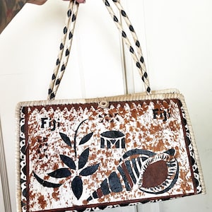 Könnte beinhalten: Eine weiße und braune gewebte Handtasche mit schwarzen Ledergriffen. Die Tasche hat ein braunes und weißes Muster mit dem Wort "Fiji" darauf.