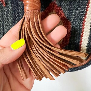 K&ouml;nnte beinhalten: Nahaufnahme eines braunen Leder-Quastens mit einem gewebten Muster. Der Quasten ist an einem gewebten Stoff mit blauen, roten und wei&szlig;en Streifen befestigt.