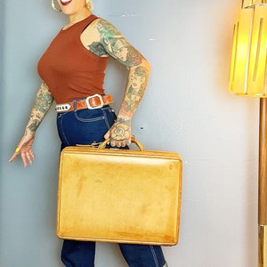 May include: A woman wearing a rust-colored tank top and blue jeans is holding a tan leather suitcase with a metal handle.