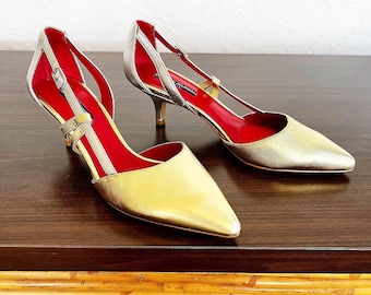 Puede incluir: Un par de zapatos de tacón dorados y plateados con forro rojo y una correa en el tobillo. Los zapatos tienen una punta puntiaguda y un tacón bajo.