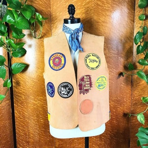 May include: A tan leather vest with various patches, including "Indian Guides", "Puma Nation", "Maria T.H.C.A. Junior Delish Campout", and "County YMCA Pinewood Derby".