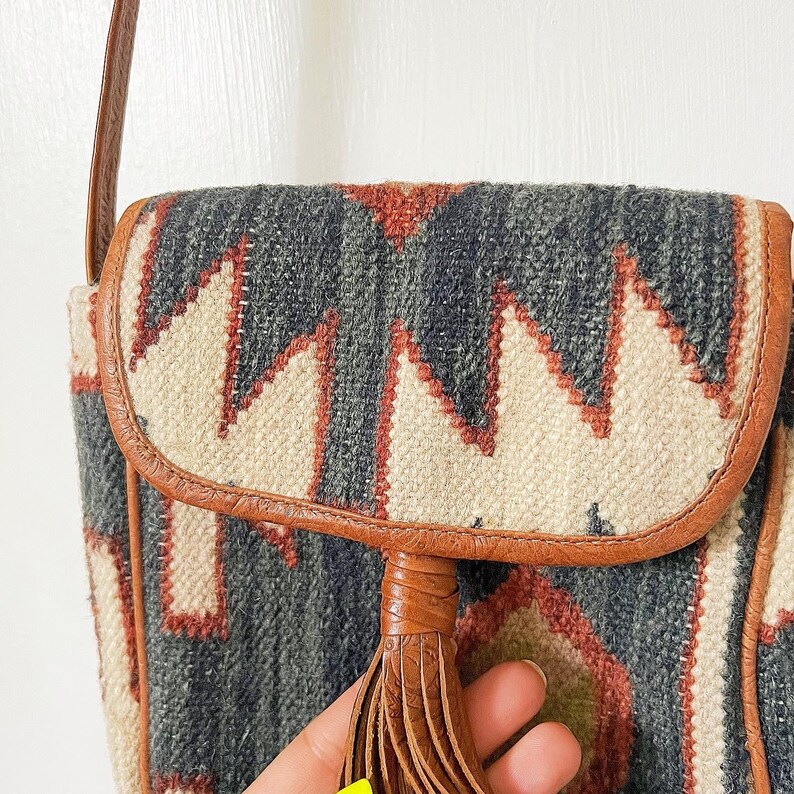 K&ouml;nnte beinhalten: Eine braune Leder-Umh&auml;ngetasche mit einem geometrischen Muster in Grau-, Wei&szlig;- und Rott&ouml;nen. Die Tasche hat einen Quastenanh&auml;nger und einen Klappenverschluss.