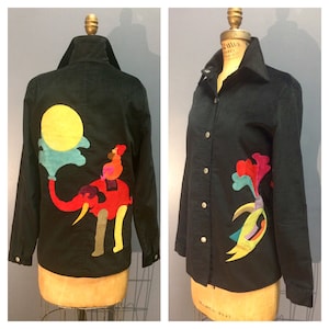 May include: A black corduroy shirt with a colorful appliqu&eacute; of an elephant, a person riding the elephant, and a sun. The appliqu&eacute; is on the back of the shirt. The shirt has a button-up front and long sleeves.