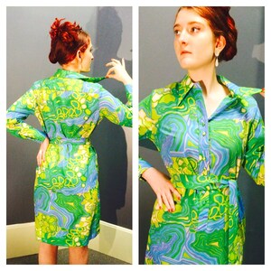 Pode incluir: Um vestido estampado verde, azul e branco com gola e mangas compridas. O vestido tem um cinto e um fecho de bot&atilde;o.