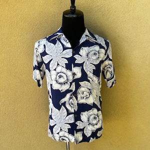 Puede incluir: Una camisa de manga corta azul marino con estampado floral blanco. Las flores son estilizadas y se asemejan a flores de hibisco. La camisa tiene un frente abotonado y un cuello.