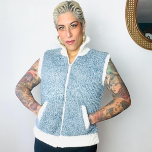 May include: A blue and white knitted vest with a zip closure and two pockets. The vest has a white trim around the neckline and the bottom.