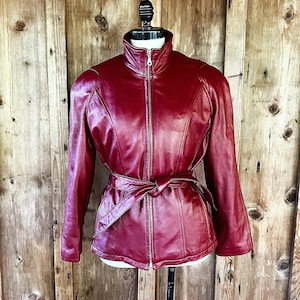 May include: A burgundy leather jacket with a zipper closure and a belt. The jacket has a stand-up collar and long sleeves.