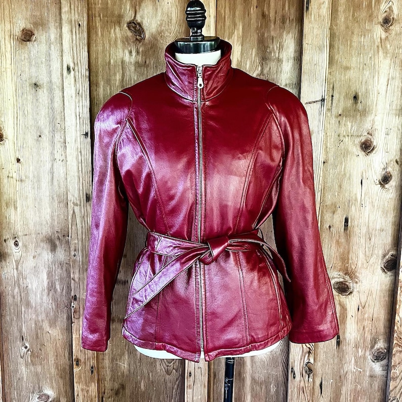 May include: A burgundy leather jacket with a zipper closure and a belt. The jacket has a stand-up collar and long sleeves.