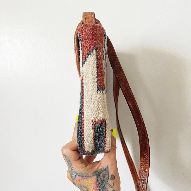 K&ouml;nnte beinhalten: Eine kleine, wei&szlig;e und braune Umh&auml;ngetasche mit einem braunen Lederriemen. Die Tasche hat ein geometrisches Design und ist aus gewebtem Stoff.