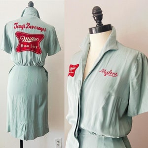 Puede incluir: Un vestido vintage azul claro con mangas cortas y cierre de botones en la parte delantera. El vestido tiene un logotipo de Miller High Life rojo y blanco en la parte posterior y el texto "Tony's Beverages" encima. La parte delantera del vestido tiene un logotipo de Miller High Life rojo y blanco en el lado izquierdo y el texto "Marlene" en el lado derecho.