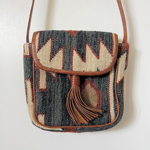 K&ouml;nnte beinhalten: Eine kleine braune Leder-Umh&auml;ngetasche mit geometrischem Muster und Quaste. Die Tasche hat einen Klappverschluss und einen langen Riemen.