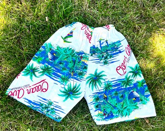 May include: White swim trunks with a blue, green, and red tropical print. The trunks feature palm trees, a sailboat, and the text "Ocean Air" with the words "Since 1997" underneath.