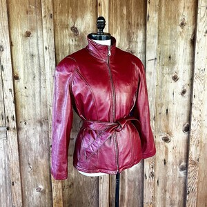 May include: A red leather jacket with a zipper closure and a belt. The jacket has a stand-up collar and long sleeves.