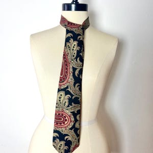 Puede incluir: Una corbata negra con estampado paisley con detalles en rojo, dorado y verde. La corbata está atada alrededor del cuello de un maniquí.