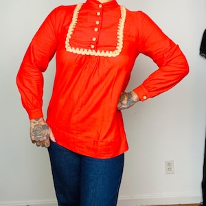 Può includere: Una camicetta rossa brillante a maniche lunghe con colletto alto e un bordo smerlato color crema intorno allo scollo. La camicetta ha piccoli bottoni rotondi color crema sul davanti e sui polsini. La persona indossa jeans blu scuro.