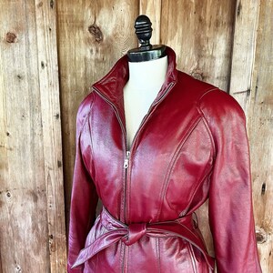 May include: A red leather jacket with a belted waist and a zipper closure. The jacket has a stand-up collar and long sleeves.