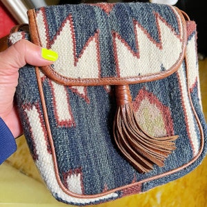 K&ouml;nnte beinhalten: Eine kleine braune Leder-Umh&auml;ngetasche mit einem geometrischen Muster in Wei&szlig;-, Blau- und Rott&ouml;nen. Die Tasche hat einen Quastenanh&auml;nger und einen Klappenverschluss.