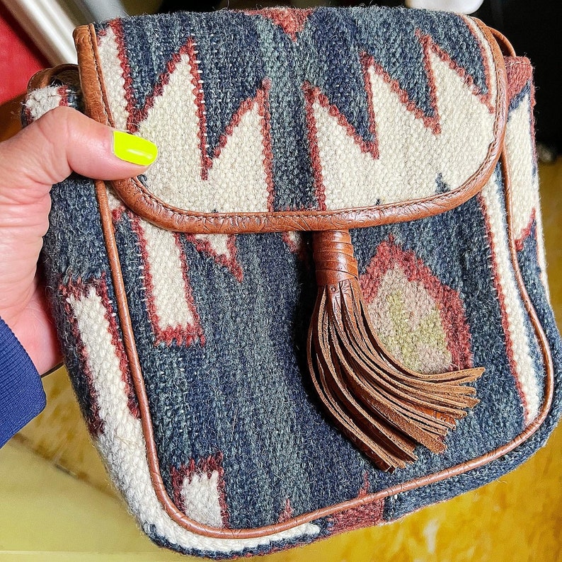 K&ouml;nnte beinhalten: Eine kleine braune Leder-Umh&auml;ngetasche mit einem geometrischen Muster in Wei&szlig;-, Blau- und Rott&ouml;nen. Die Tasche hat einen Quastenanh&auml;nger und einen Klappenverschluss.