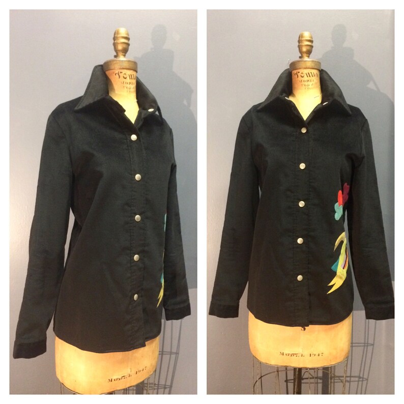 May include: A black corduroy shirt with a colorful embroidered design on the right side. The shirt has a button-up front and long sleeves.