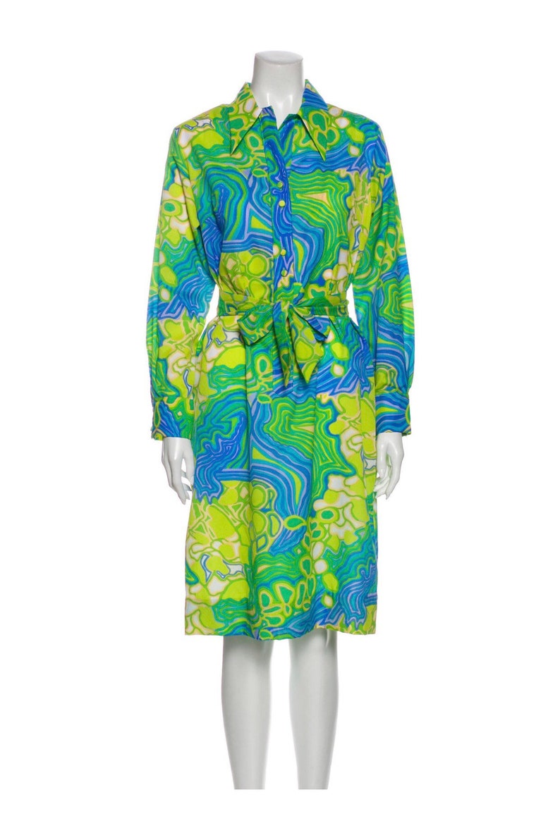 Pode incluir: Um vestido de manga comprida com estampa abstrata verde, azul e branca. O vestido tem gola, fechamento frontal com bot&otilde;es e um cinto de amarra&ccedil;&atilde;o na cintura.