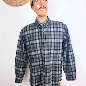 May include: A long-sleeve button-down shirt featuring a black, gray, and white plaid pattern. The shirt has a standard collar, a single chest pocket, and a buttoned front placket. The fabric has a soft, brushed texture throughout the garment.