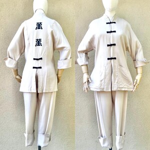 Puede incluir: Un conjunto de dos piezas en color beige claro. La chaqueta tiene cuello mandarín, mangas largas y cierre frontal con botones negros. El pantalón tiene pierna recta y dobladillo con vuelta. La chaqueta tiene caracteres negros bordados en la espalda.