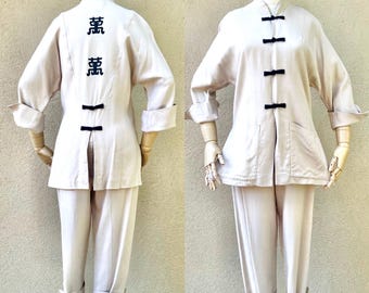 Pode incluir: Um conjunto de duas peças em cor bege claro. A jaqueta tem gola mandarim, mangas compridas e fecho frontal com botões pretos. A calça tem perna reta e bainha dobrada. A jaqueta tem caracteres pretos bordados nas costas.