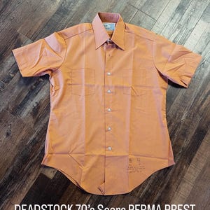 Puede incluir: Una camisa color melocotón de manga corta con dos bolsillos en el pecho y cuello puntiagudo. La camisa tiene botones frontales y está etiquetada como "DEADSTOCK 70's Sears PERMA PREST 44/46 Chest."