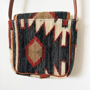 K&ouml;nnte beinhalten: Eine kleine, schwarz-wei&szlig;e Umh&auml;ngetasche mit einem braunen Lederriemen und Bes&auml;tzen. Die Tasche hat ein geometrisches Design mit roten, wei&szlig;en und braunen Akzenten.