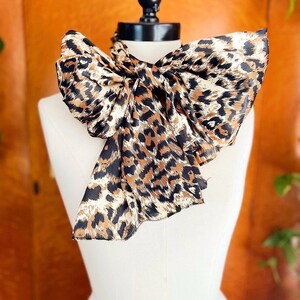 Könnte beinhalten: Ein Schal mit Leopardenmuster, zu einer großen Schleife gebunden, wird auf einer weißen Schaufensterpuppe präsentiert. Der Schal hat schwarze Flecken und braune Flecken auf cremefarbenem Hintergrund. Der Schal ist aus seidigem Material.