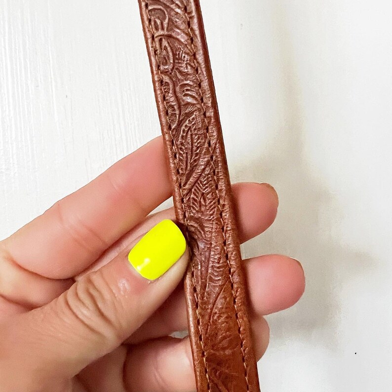 K&ouml;nnte beinhalten: Nahaufnahme eines braunen Lederriemens mit einem gepr&auml;gten Blumenmuster. Der Riemen wird in einer Hand mit gelbem Nagellack gehalten.