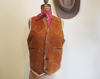 May include: A brown suede vest with a faux fur collar and a red bandana around the neck. The vest has a button closure and two pockets.