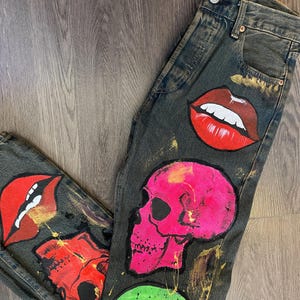 Puede incluir: Vaqueros de mezclilla gris oscuro con diseños pintados a mano que incluyen una calavera grande de color rosa brillante, una calavera verde, una calavera roja y dos pares de labios rojos con dientes blancos. Los vaqueros tienen bragueta de botones.
