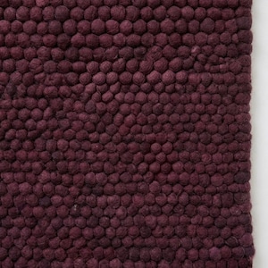 Puede incluir: Una alfombra de color ciruela intenso construida a partir de una densa disposición de pequeñas bolas de lana afieltrada esféricas. La superficie presenta un patrón texturizado uniforme creado por los elementos de fieltro redondeados.