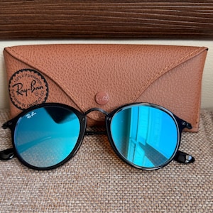 May include: Black-framed sunglasses with reflective blue lenses and the Ray-Ban logo on the upper corner of the left lens. The sunglasses are positioned in front of a tan, textured, triangular-shaped protective case with a Ray-Ban logo stamp.