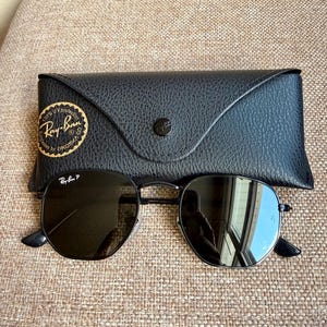 May include: Black Ray-Ban sunglasses with hexagonal frames and dark polarized lenses, featuring the Ray-Ban P logo on the lens. The sunglasses are paired with a black textured leather-like carrying case with a gold circular seal.