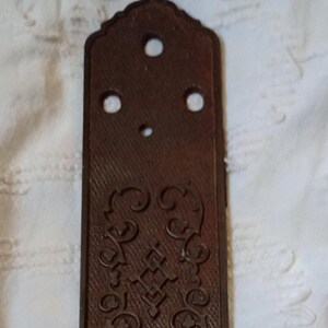 May include: A rectangular, antique brown metal door plate with an ornate, embossed design. The plate features a textured surface, decorative scrollwork, and several holes for mounting. The overall design is symmetrical.